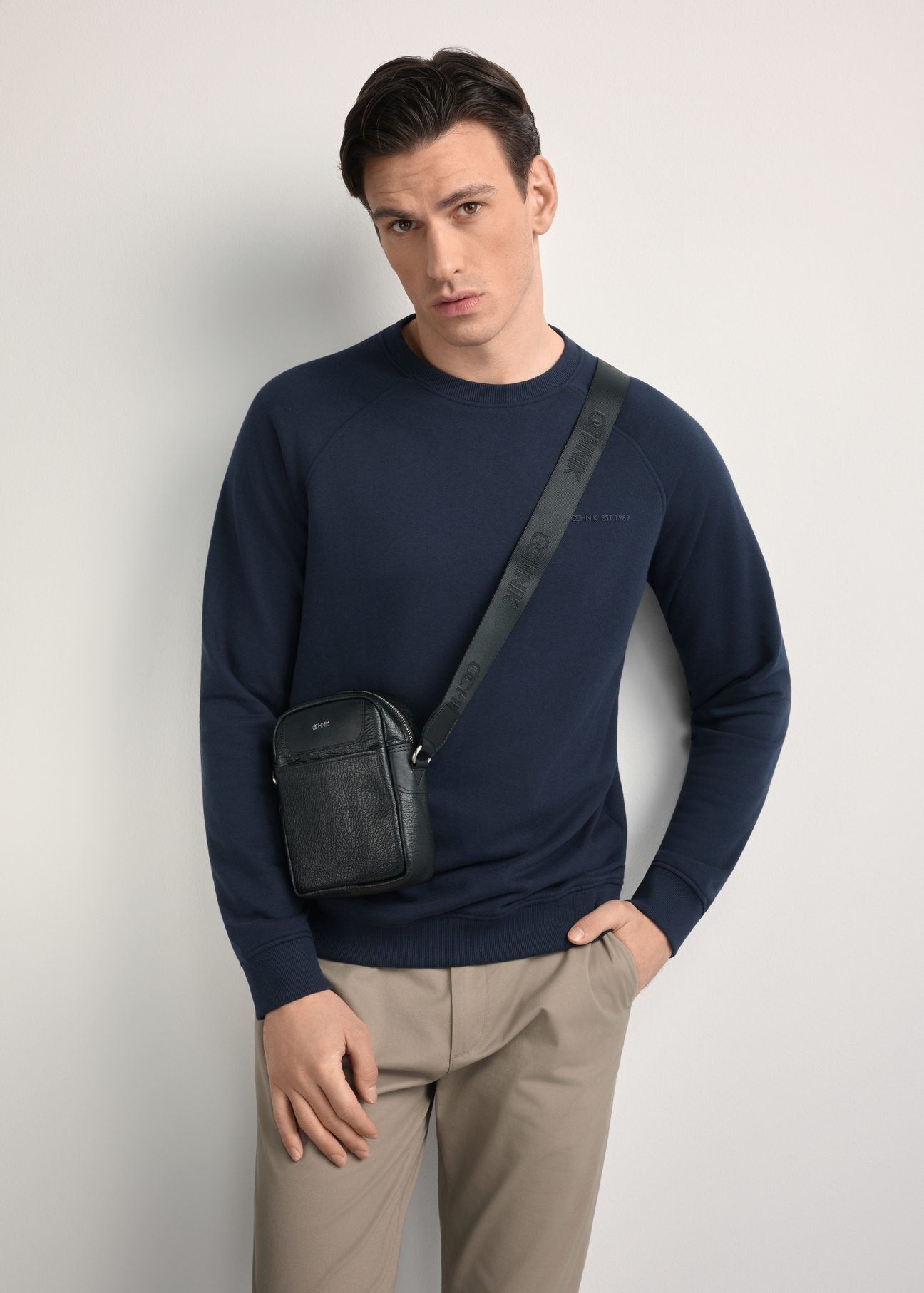 Men's black leather waist bag with TORMS-0296A-99(Z25) logo - diagonal product view, modeled on a white background. Knee-length silhouette visible.